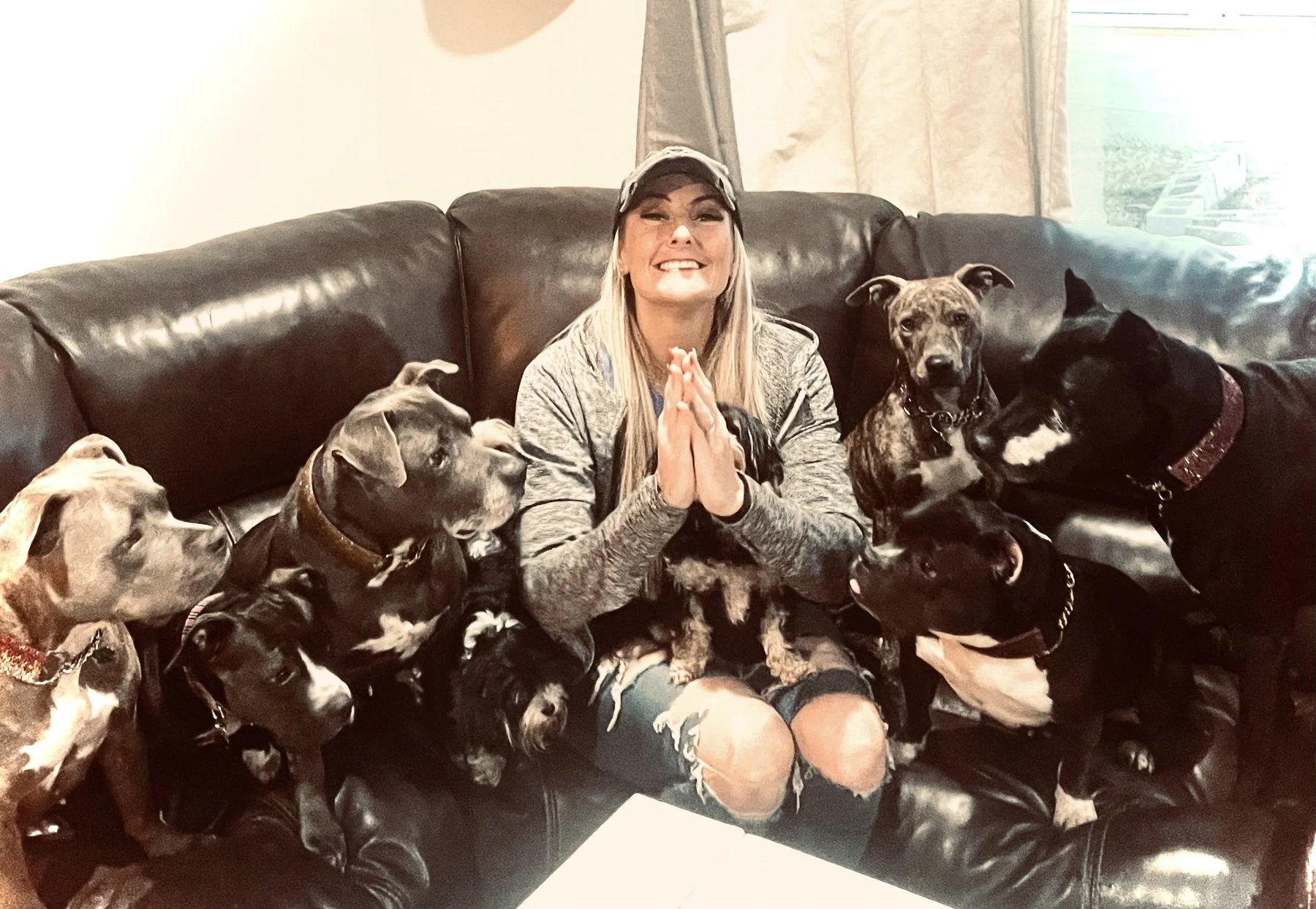 Tiffany Skuraton, founder of Blue Waters Dog Rescue, smiling on a leather couch with hands pressed together, surrounded by rescue dogs